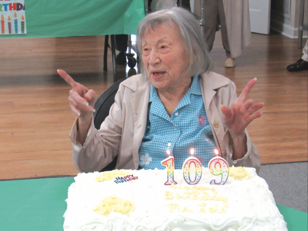 Cook County's oldest citizen, Mrs. Lois Musselwhite is pictured wearing a blue button up with a tan jacket. She is sitting at a table with a green tablecloth and a birthday cake with lit candles. The rainbow candles read out, "109."