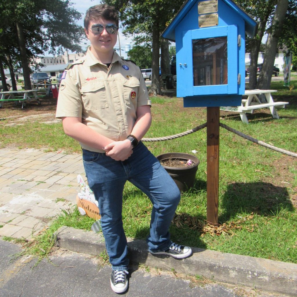 Boy Scout David M. Greene stands to the left of a vibrant blue Little Library clasping his hands in front of him. He is wearing his Boy Scouts shirt, aviator sunglasses, and blue jeans. The blue little library is placed outside of the Cook County library