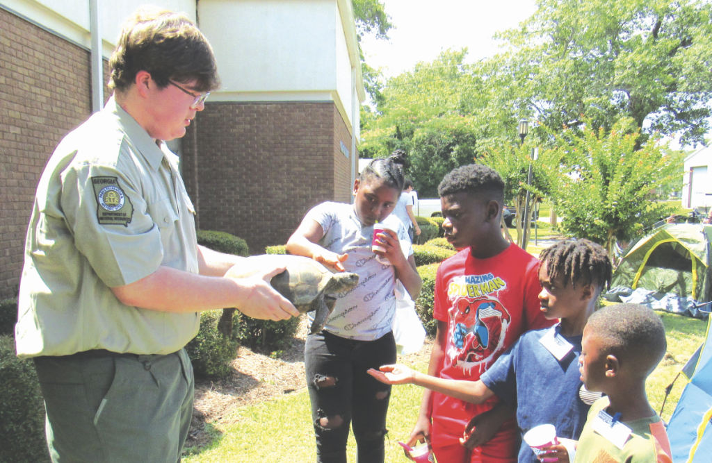 A Park Ranger from Reed Bingham State parks holds a tortoise for 4 children with outstreched hands, reaching to pet it.