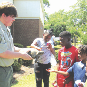 A Park Ranger from Reed Bingham State parks holds a tortoise for 4 children with outstreched hands, reaching to pet it.