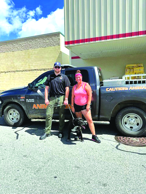 Animal control officer Ronan Ward stands on the left holding a black leashed dog. Next to him on the right is Kylie Evans wearing a hot pink tank top and shorts. They are standing in front of an animal control truck.