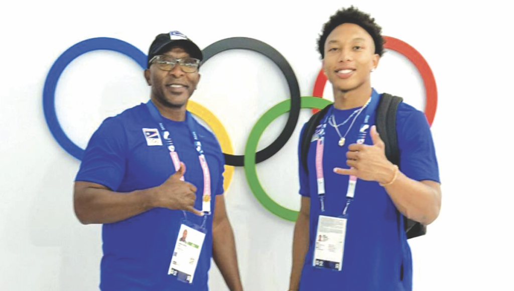 Geary Reed and his son William (Will) are shown wearing blue polo shirts. They are shown doing the shaka ("hang loose") sign in front of the logo of Olympic rings on a white wall. 
