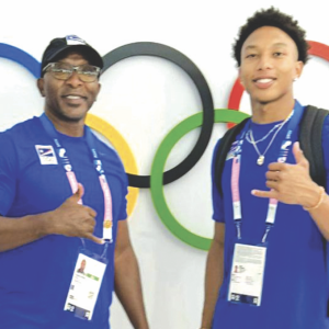 Geary Reed and his son William (Will) are shown wearing blue polo shirts. They are shown doing the shaka ("hang loose") sign in front of the logo of Olympic rings on a white wall.