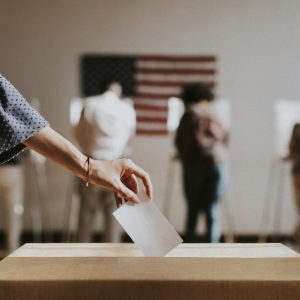 a woman's hand drops a vote in a ballot box. in the background there are people voting on machines and an american flag hanging on a white wall